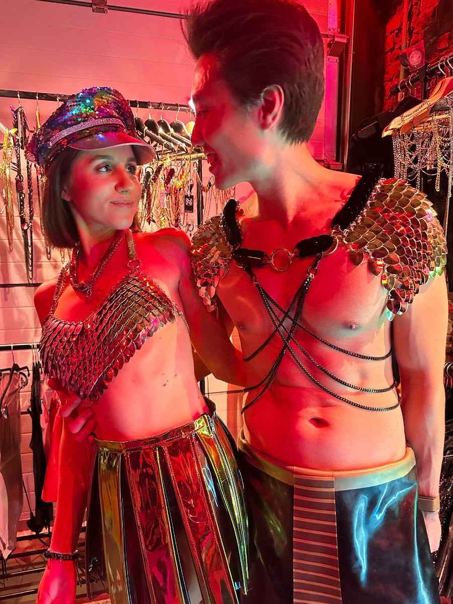 Couple wearing silver metallic body chains and harnesses with scale designs, colorful reflective skirts, smiling under red club lighting