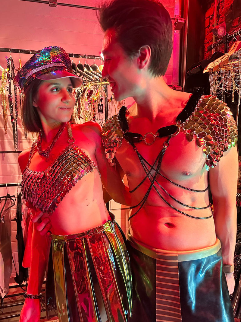 Couple wearing silver metallic body chains and harnesses with scale designs, colorful reflective skirts, smiling under red club lighting