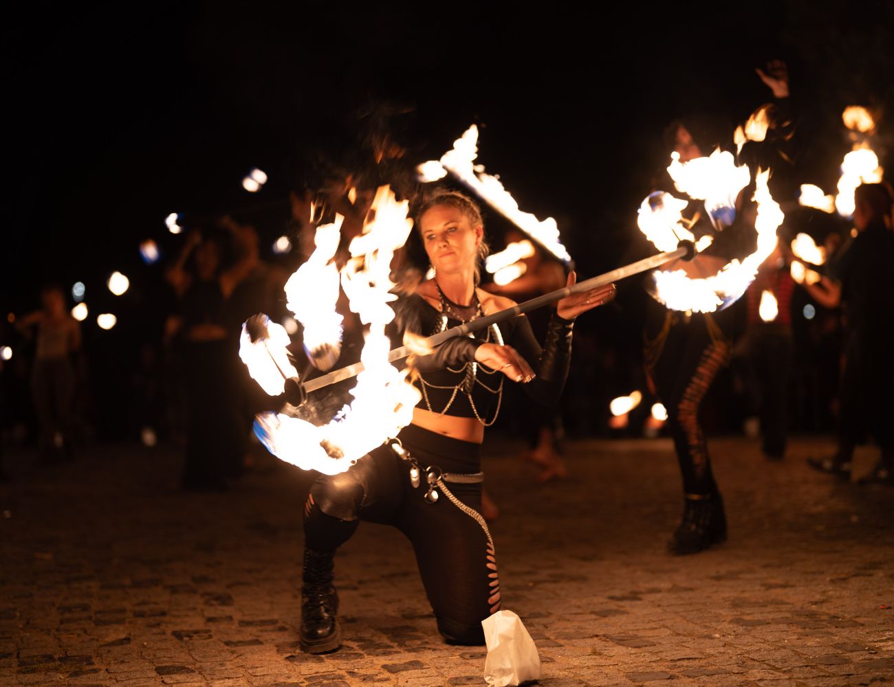 Fire performer wearing silver body chains in black outfit, dancing with flaming staffs, focused and intense