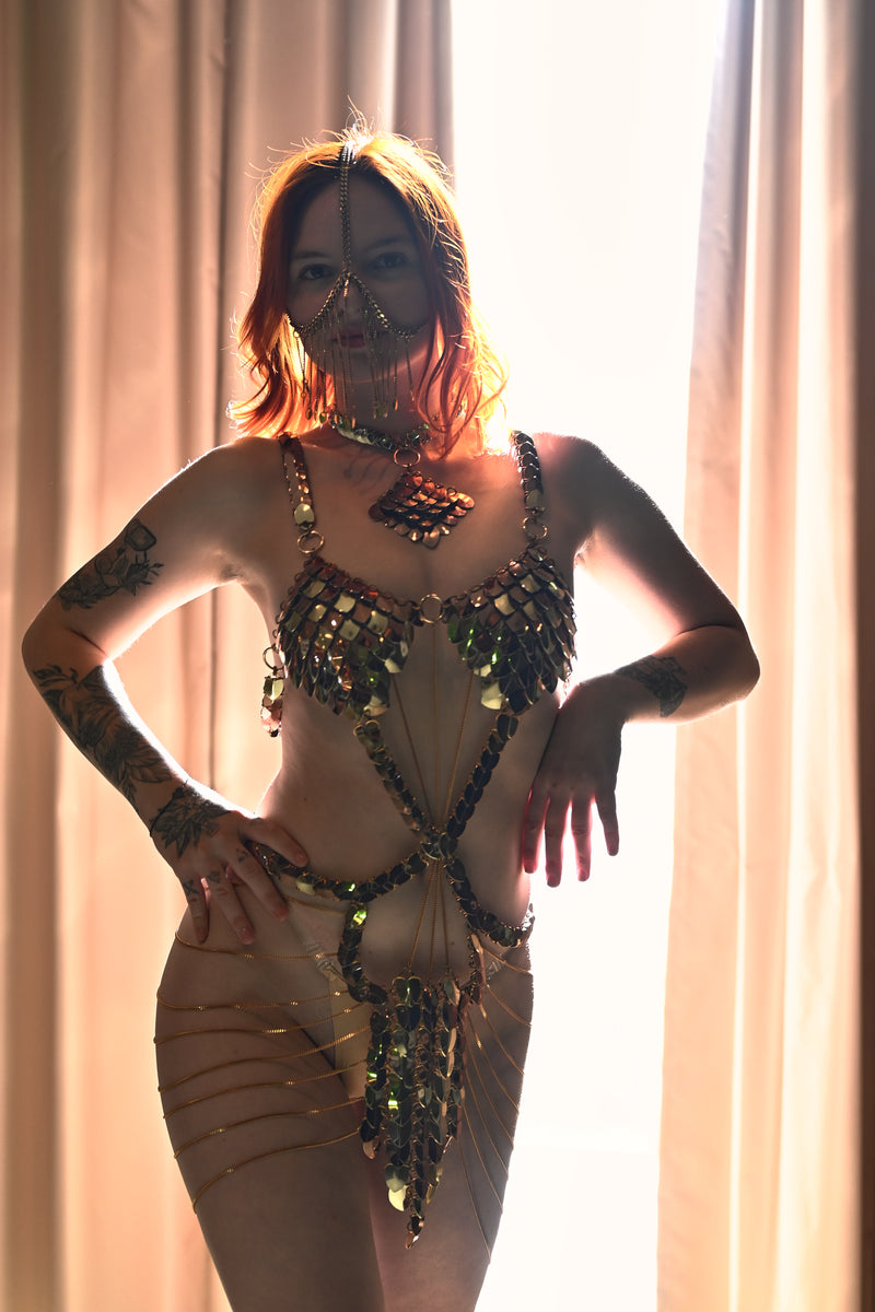 Gold chain body harness with mask worn by confident tattooed redhead woman in soft backlight
