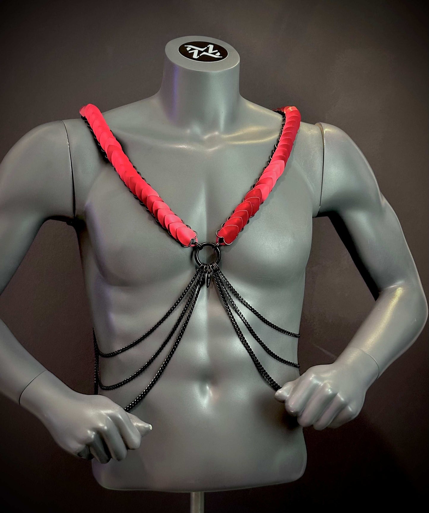 Harness mit schwarzen Ketten und roten Plättchen, stylisch und provokativ am grauen Torso