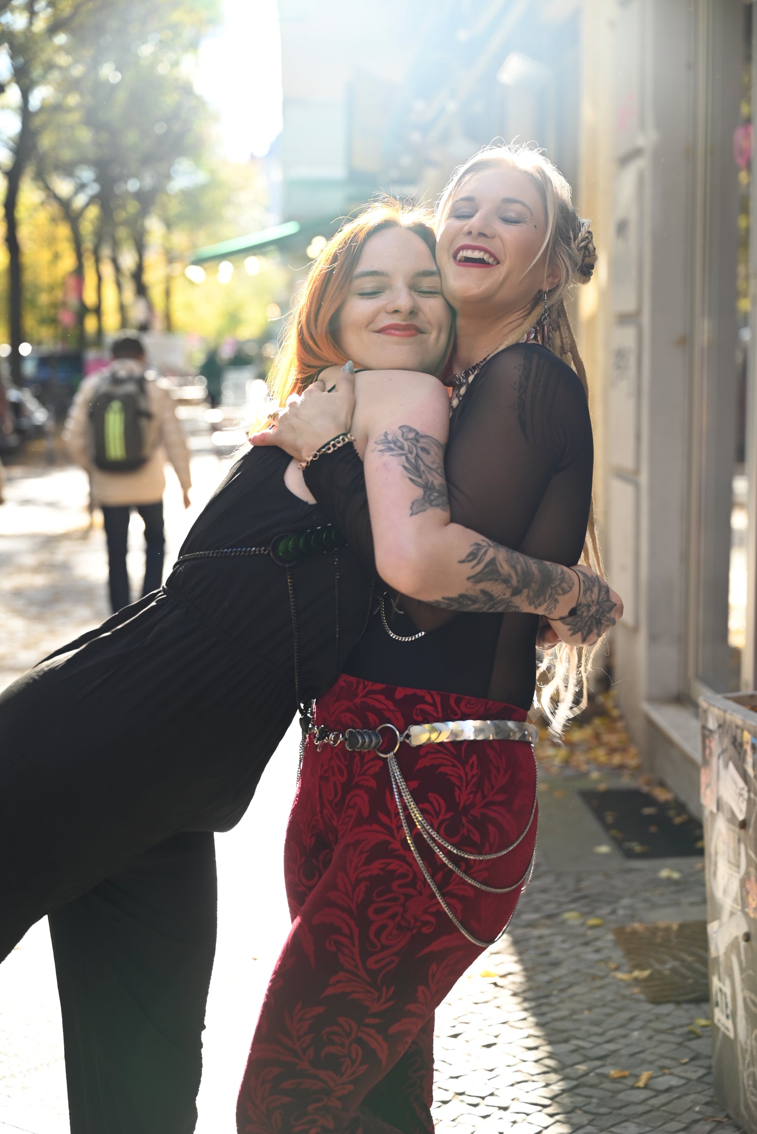 Two young women embracing on sunny city street, one in black dress, other in red patterned pants, joyful
