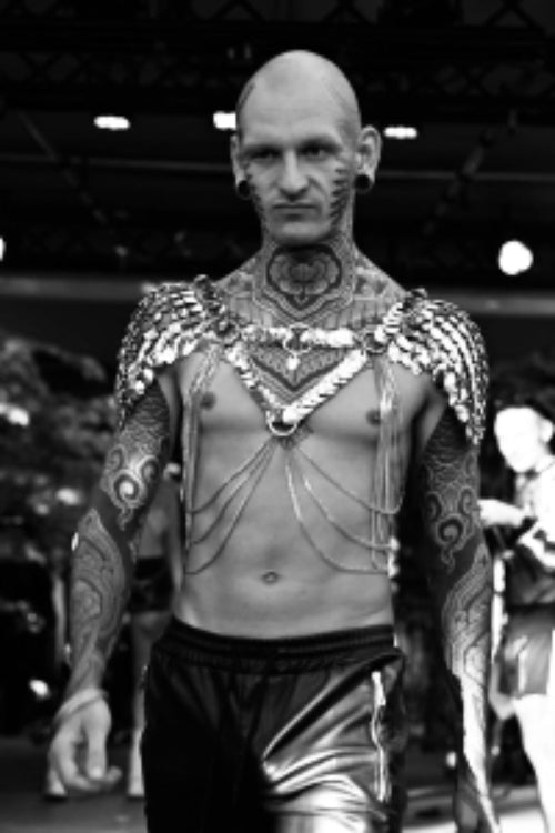 tattooed man wearing metallic chain shoulder accessory and shiny bottoms, black and white fashion style, intense emotion